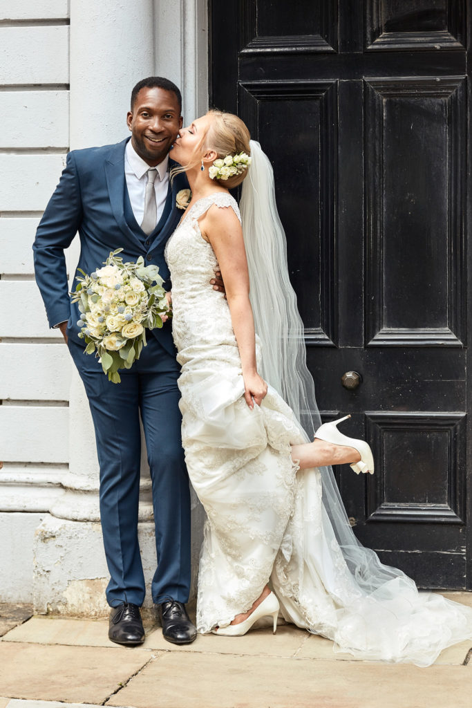 Bride kissing the groom's cheek with one foot kicked up in front of a black door